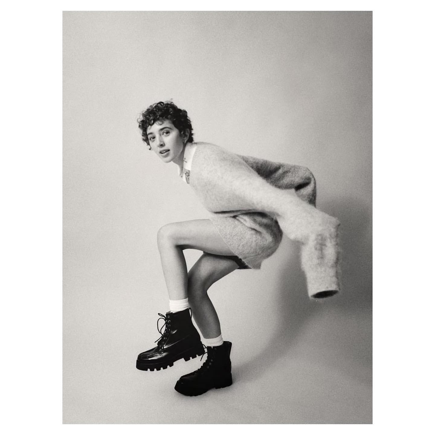 Black and white shot of a woman in a sweater and boots crouching, looking at the camera, set against a light backdrop.
