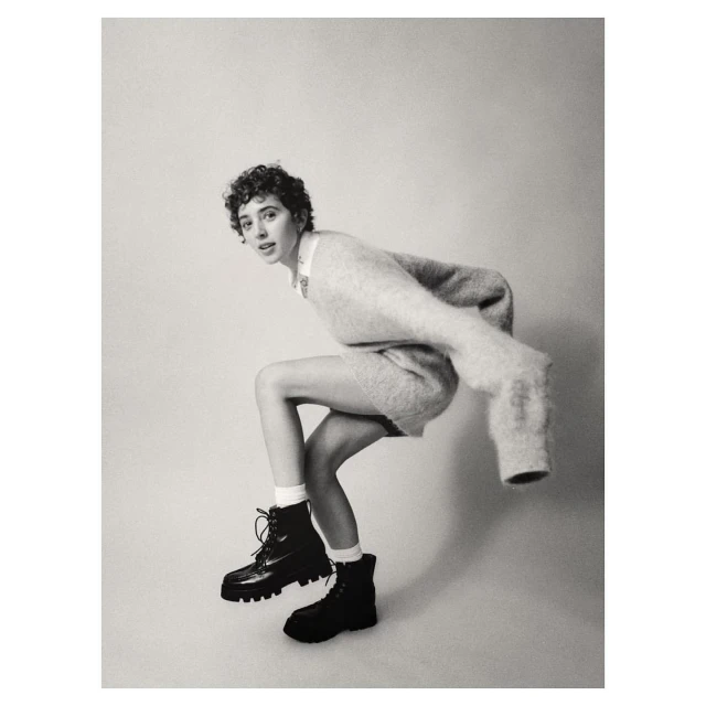 Black and white shot of a woman in a sweater and boots crouching, looking at the camera, set against a light backdrop.