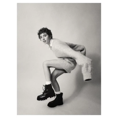 Black and white shot of a woman in a sweater and boots crouching, looking at the camera, set against a light backdrop.