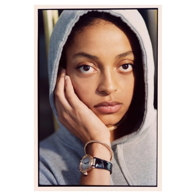 Close-up of a young Black woman wearing a gray hoodie, with a rose gold watch and beaded bracelet, resting her chin on her hand.