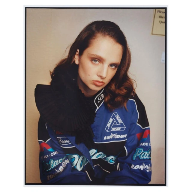 Young woman in blue Palace London racing jacket with black pleated ruff collar, looking at the camera in a studio setting.