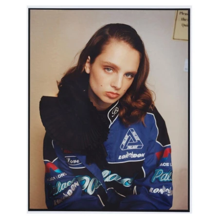 Young woman in blue Palace London racing jacket with black pleated ruff collar, looking at the camera in a studio setting.