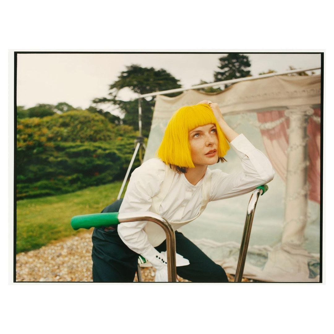 Woman in yellow wig poses outdoors on metal frame. Shirt, dark pants, trees, lawn, tent backdrop. Fashion shot, looking to upper right.