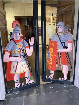 Cartoon-style Roman soldiers adorn glass doors, one waving with a spear and shield, creating a playful and historical entrance.