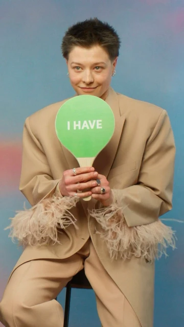 Woman in a beige suit sits on a stool, holding a green sign with the words 'I HAVE'. Studio shot with a vibrant blue and pink background.