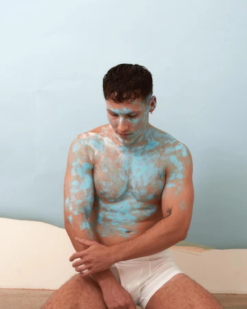Shirtless man with blue paint smeared across his torso and face wearing white briefs against a light blue backdrop.
