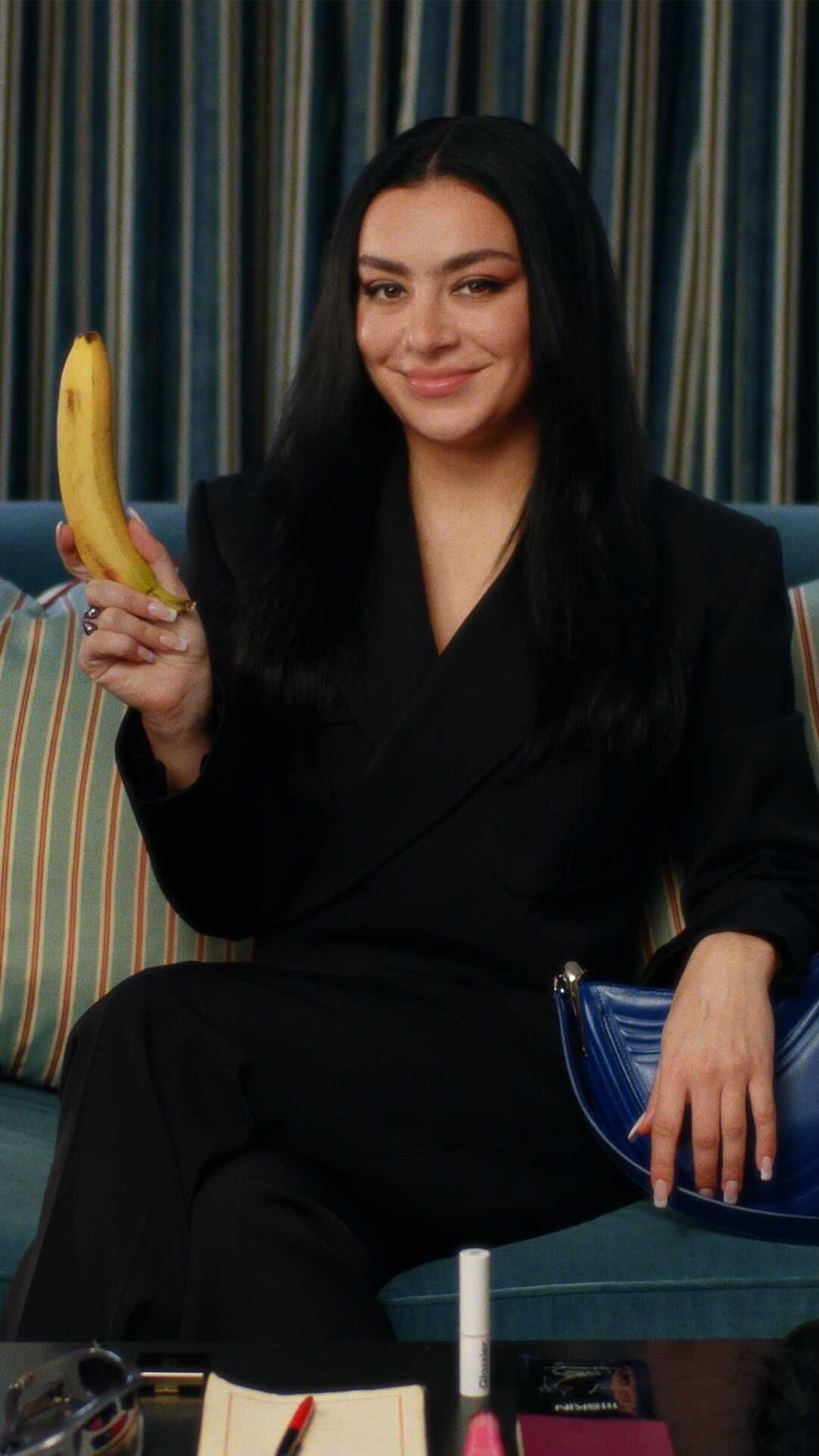 A brunette adult female in a black suit sits holding a banana, indoors. Other objects include a purse and makeup on a coffee table.