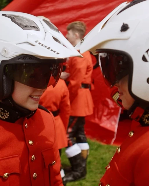 Two people wearing motorcycle helmets and red jackets face each other in an outdoor setting with a group of people in the background.