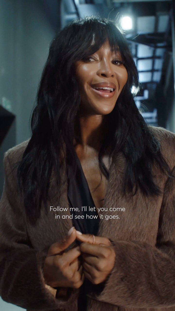 Naomi Campbell is indoors, posing in a long brown coat and looking at the camera. Dark hair with bangs frames her face.