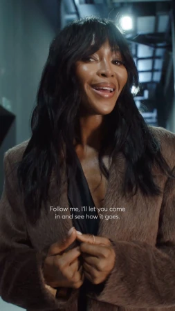 Naomi Campbell is indoors, posing in a long brown coat and looking at the camera. Dark hair with bangs frames her face.