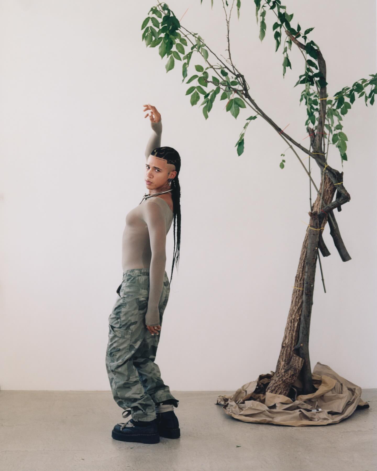 Stylish non-binary person posing next to tree with camouflage pants and grey long-sleeve top in a bright white studio setting.