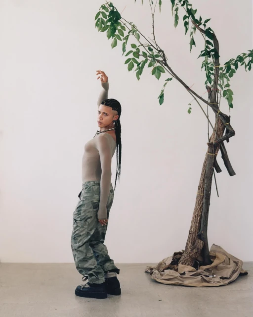 Stylish non-binary person posing next to tree with camouflage pants and grey long-sleeve top in a bright white studio setting.