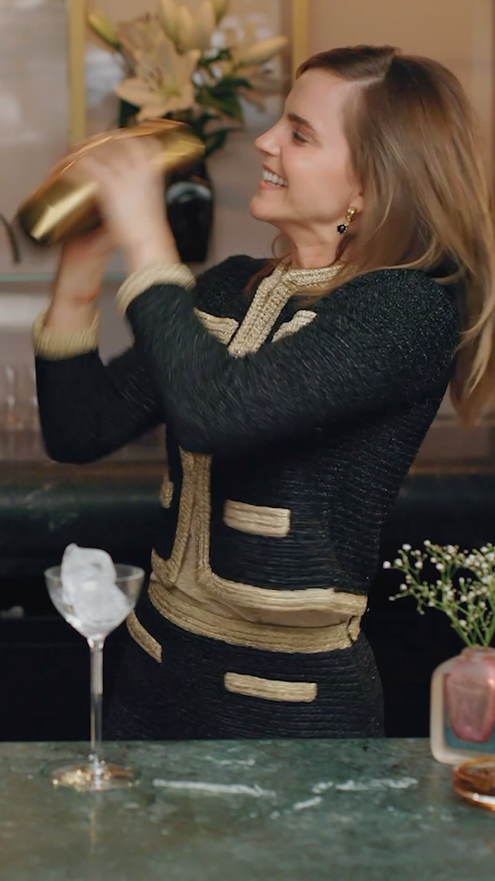 Smiling woman in black-and-gold jacket shakes a gold cocktail shaker in a bar setting, with a martini glass of ice on the counter.