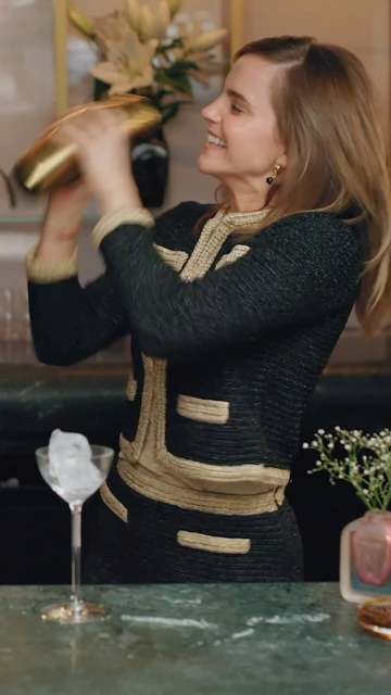 Smiling woman in black-and-gold jacket shakes a gold cocktail shaker in a bar setting, with a martini glass of ice on the counter.
