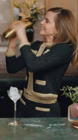 Smiling woman in black-and-gold jacket shakes a gold cocktail shaker in a bar setting, with a martini glass of ice on the counter.