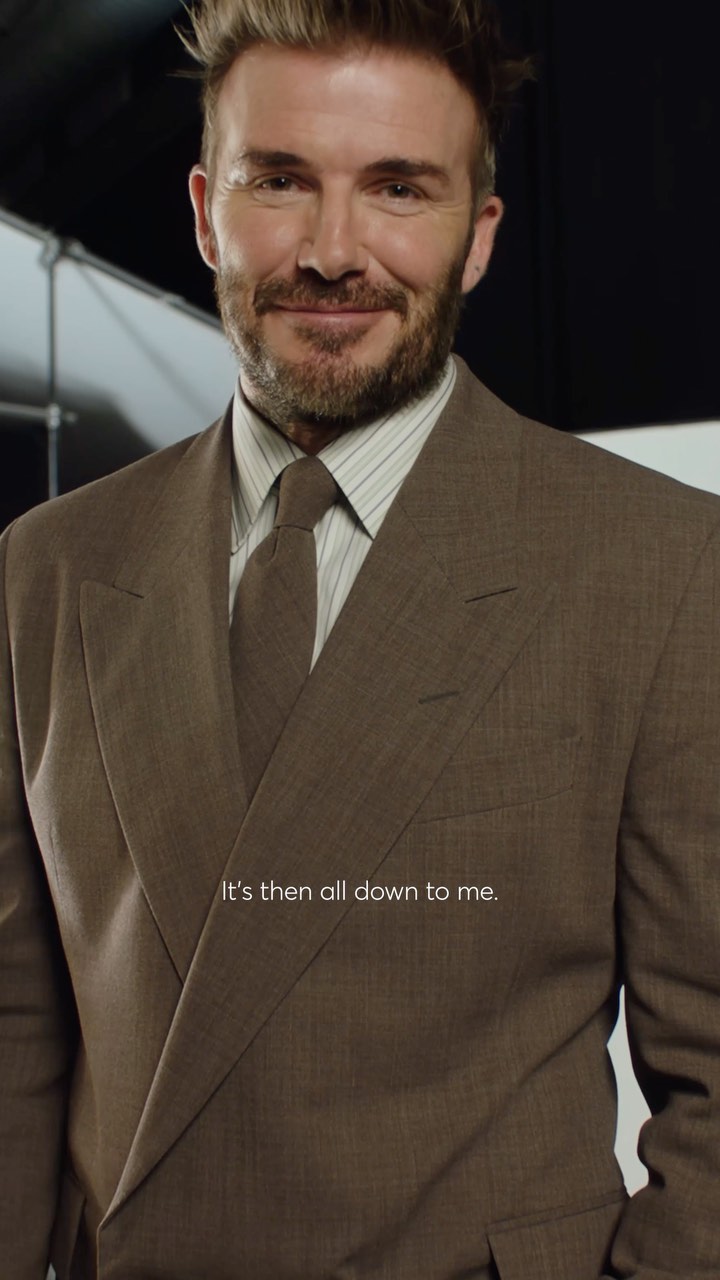 Portrait of David Beckham in a brown suit and tie with a faint smile against a softly lit background, showcasing fashion and celebrity.
