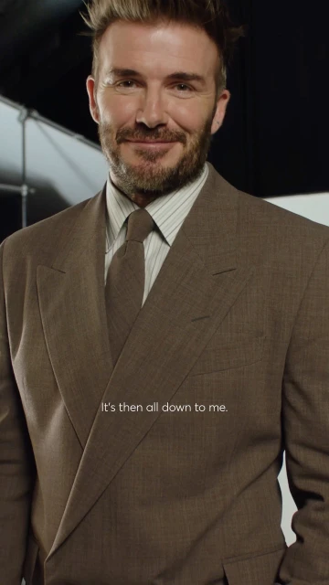 Portrait of David Beckham in a brown suit and tie with a faint smile against a softly lit background, showcasing fashion and celebrity.