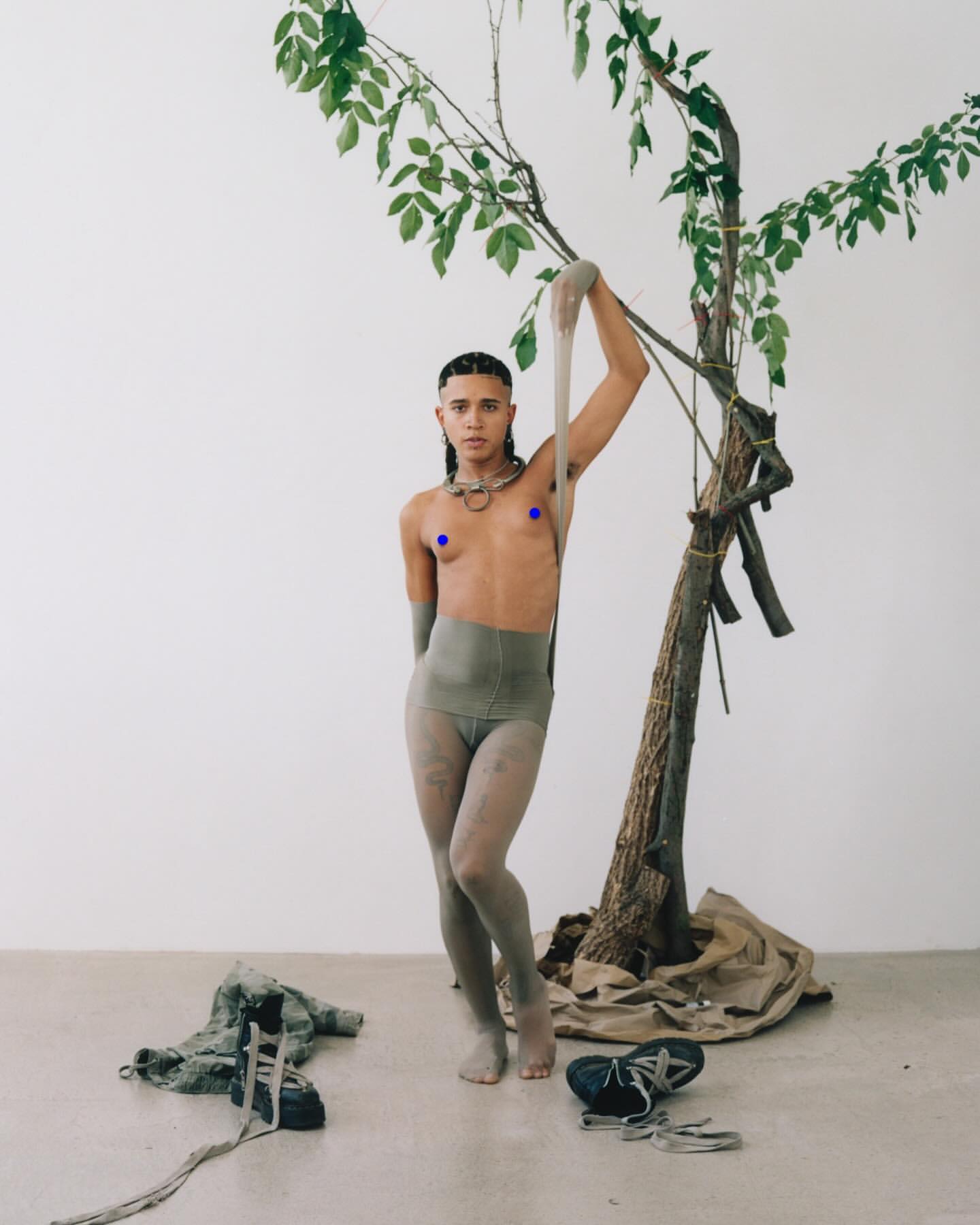 A person poses barefoot in tights and neutral-toned clothing, holding a sheer stocking, next to a small tree against a neutral background.