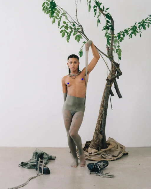 A person poses barefoot in tights and neutral-toned clothing, holding a sheer stocking, next to a small tree against a neutral background.