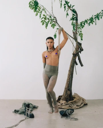 A person poses barefoot in tights and neutral-toned clothing, holding a sheer stocking, next to a small tree against a neutral background.