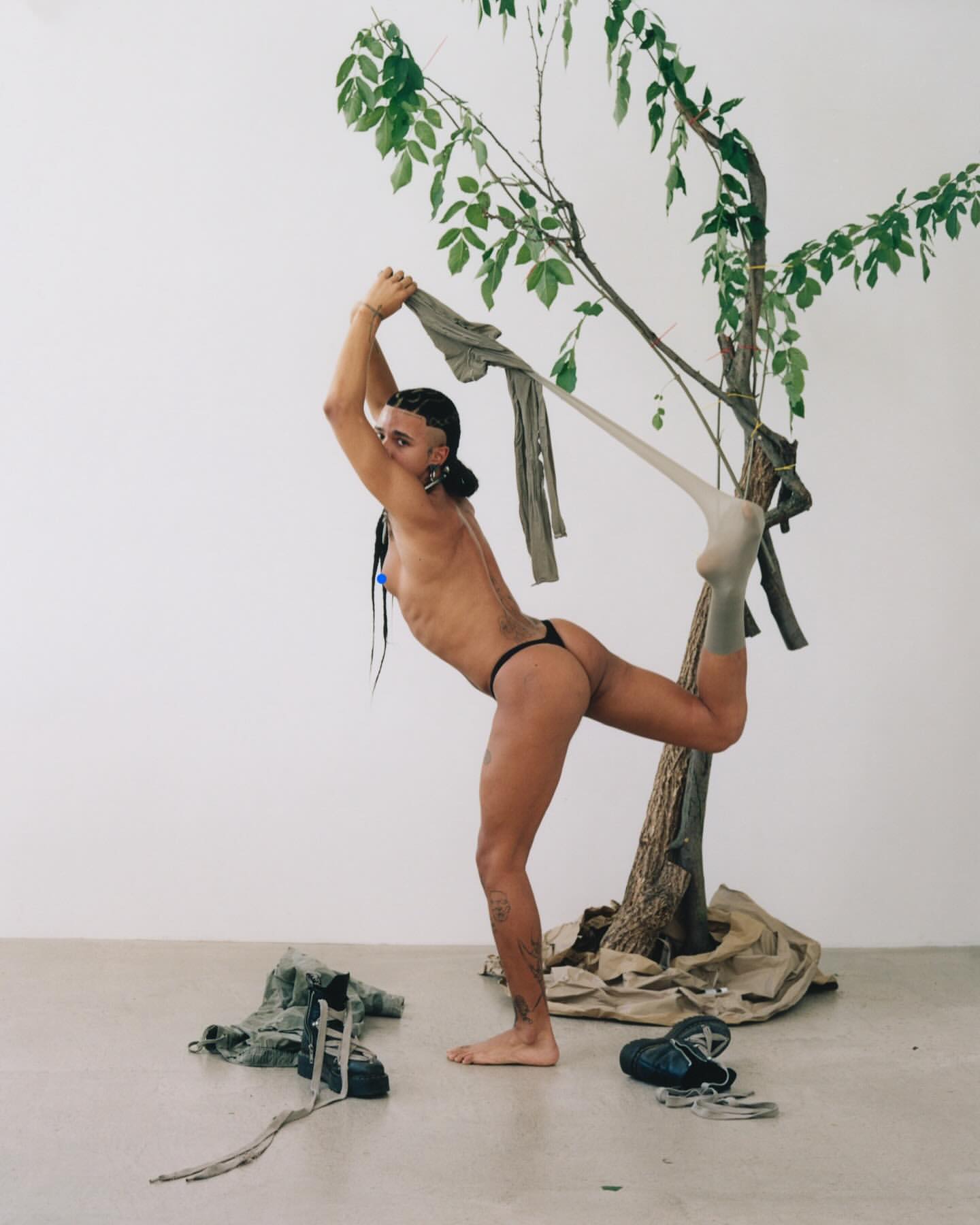 A person in a minimal outfit poses with a tree branch and olive garments in an indoor setting with bright lighting and minimal backdrop.