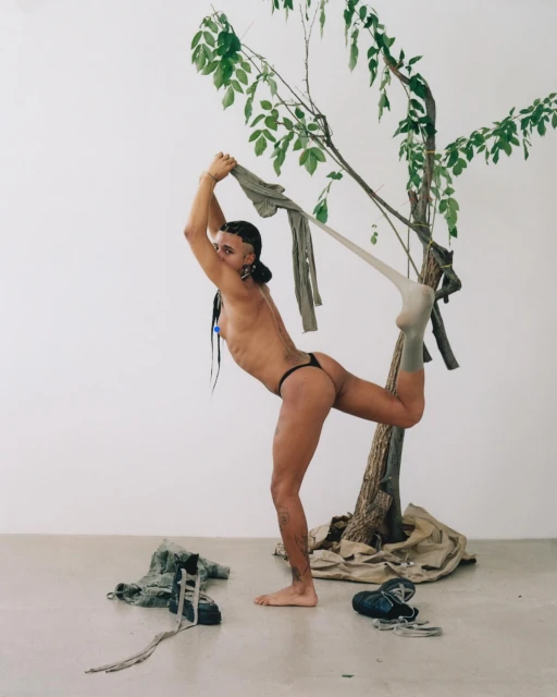 A person in a minimal outfit poses with a tree branch and olive garments in an indoor setting with bright lighting and minimal backdrop.