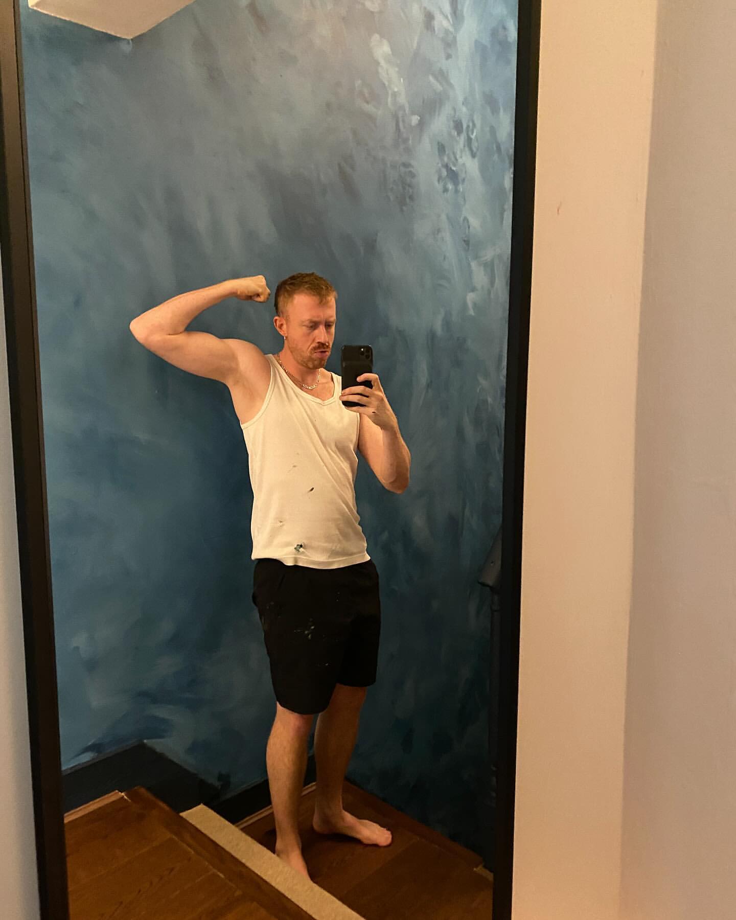 Adult male flexing muscles in a mirror wearing a tank top and black shorts with a blue painted wall in an indoor setting.
