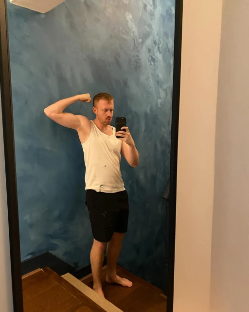 Adult male flexing muscles in a mirror wearing a tank top and black shorts with a blue painted wall in an indoor setting.