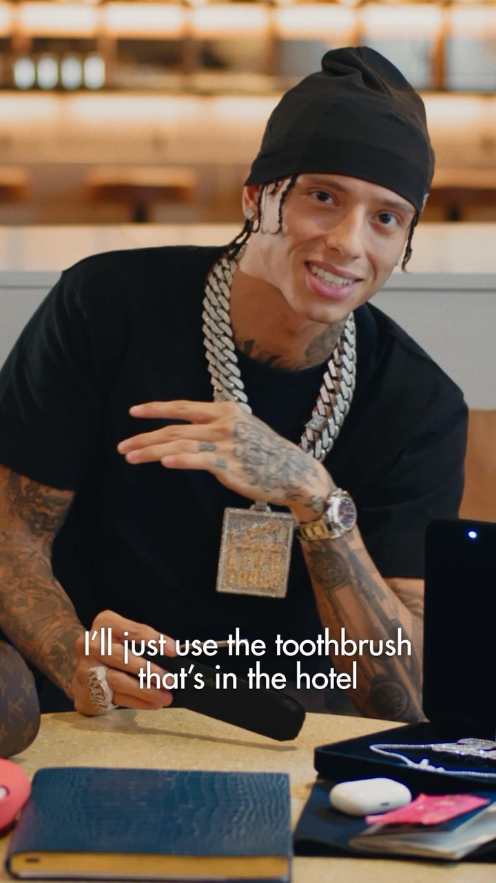 A man with a beanie and heavy jewelry sitting at a table indoors, holding a black toothbrush case. He is looking at the camera.