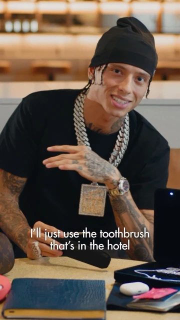 A man with a beanie and heavy jewelry sitting at a table indoors, holding a black toothbrush case. He is looking at the camera.