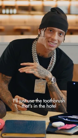 A man with a beanie and heavy jewelry sitting at a table indoors, holding a black toothbrush case. He is looking at the camera.