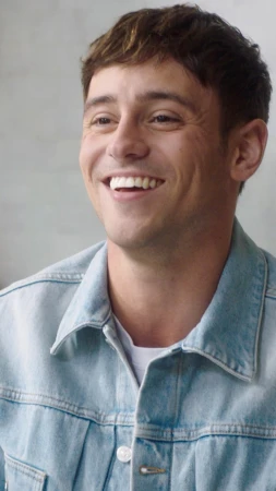 Portrait of a smiling young man wearing a light wash denim jacket over a white t-shirt against a plain neutral background.