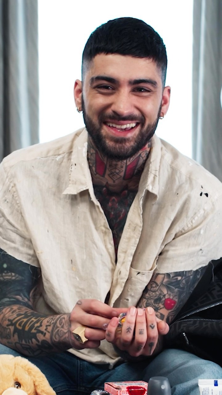 Smiling man with tattoos in an off-white shirt and blue jeans sitting indoors with scattered objects in the foreground.