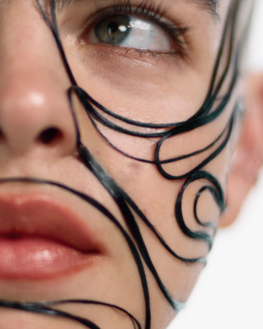 Close-up portrait of an adult woman with artistic black designs painted on her face, featuring visible green eye and partial view of lips.