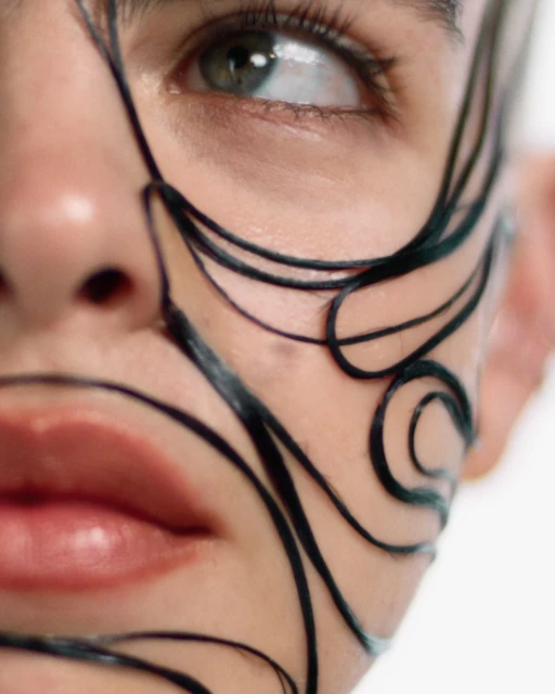 Close-up portrait of an adult woman with artistic black designs painted on her face, featuring visible green eye and partial view of lips.