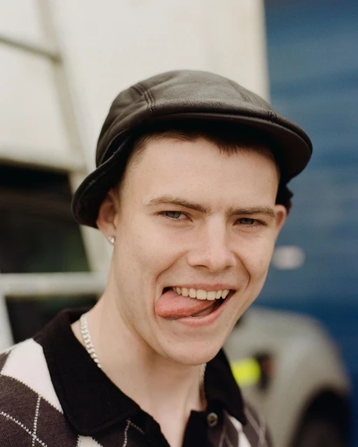 Close-up portrait of a young adult man wearing a hat and patterned shirt, sticking out his tongue and smiling in an outdoor setting.