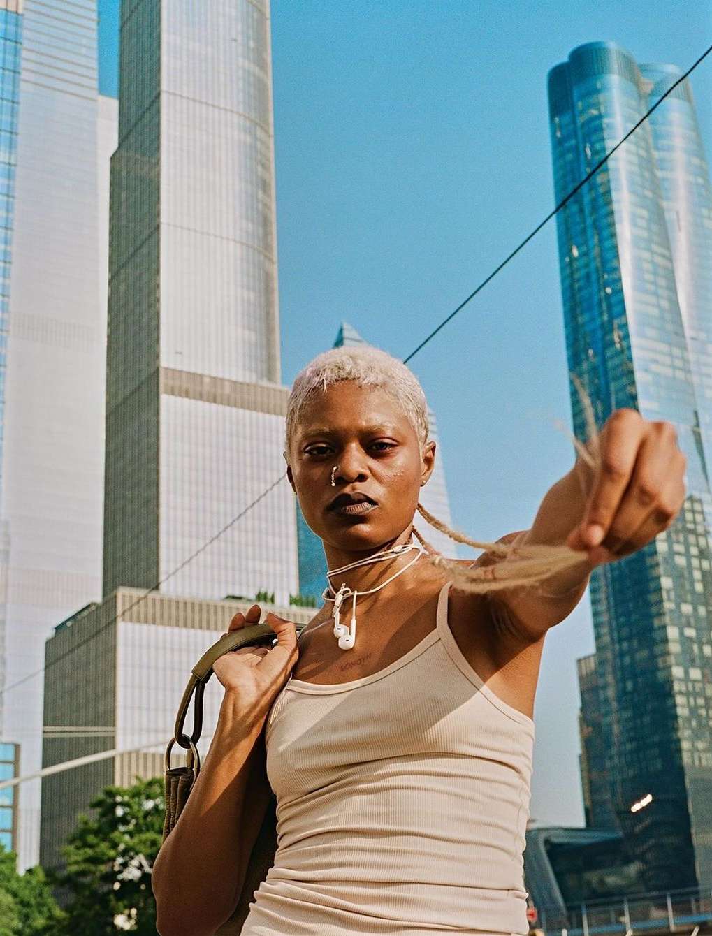 Portrait of a woman with short blonde hair in a beige tank top and light makeup standing in front of skyscrapers and a blue sky.