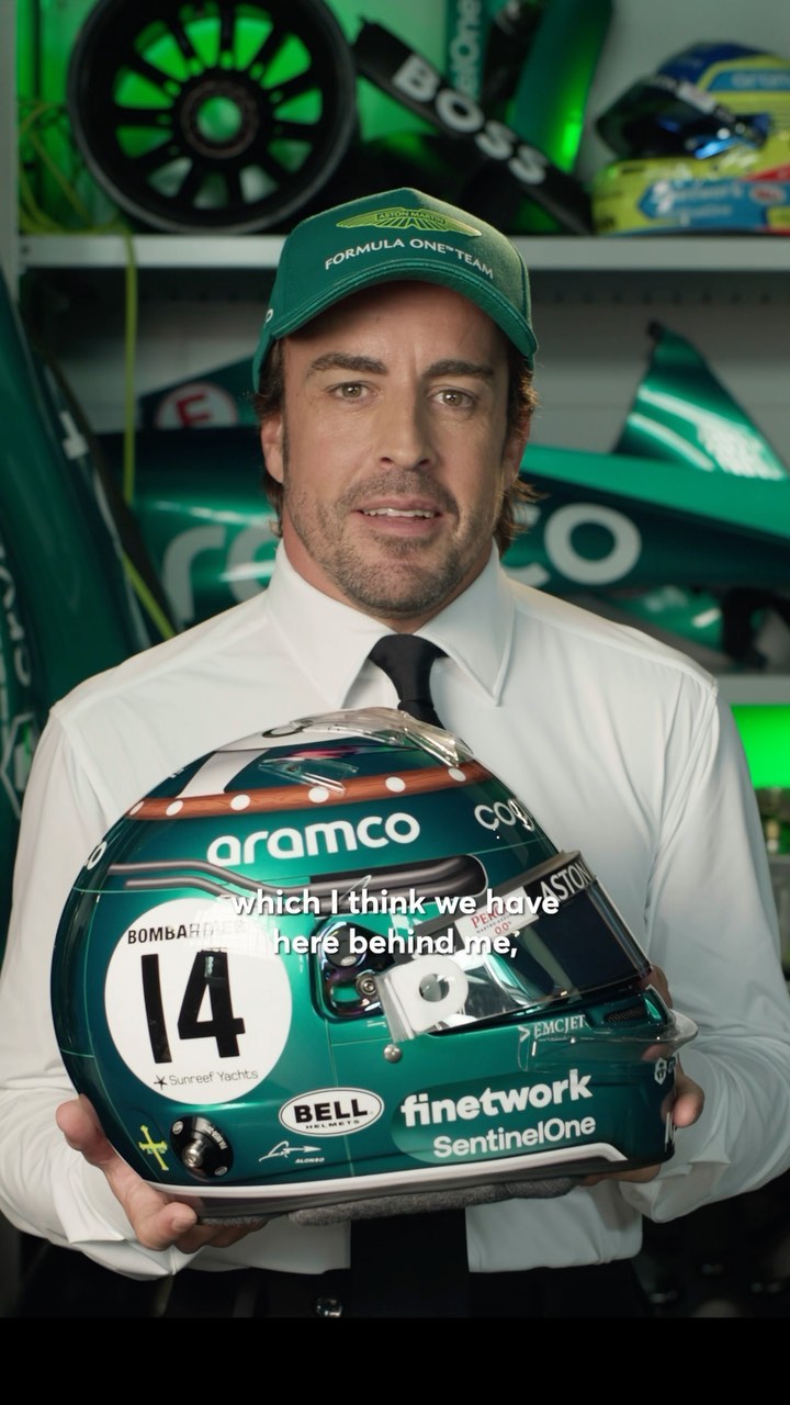 Adult male wearing Formula One team hat and a white shirt holding a racing helmet with logos and the number 14. 