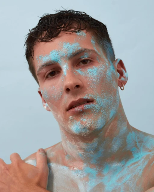 Close-up portrait of a male model with light brown hair and turquoise glitter on his face and upper body against a light blue background.