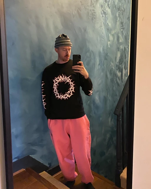 An adult male takes a selfie in a mirror wearing a beanie, black long-sleeve shirt with white graphics, and pink sweatpants on a staircase.