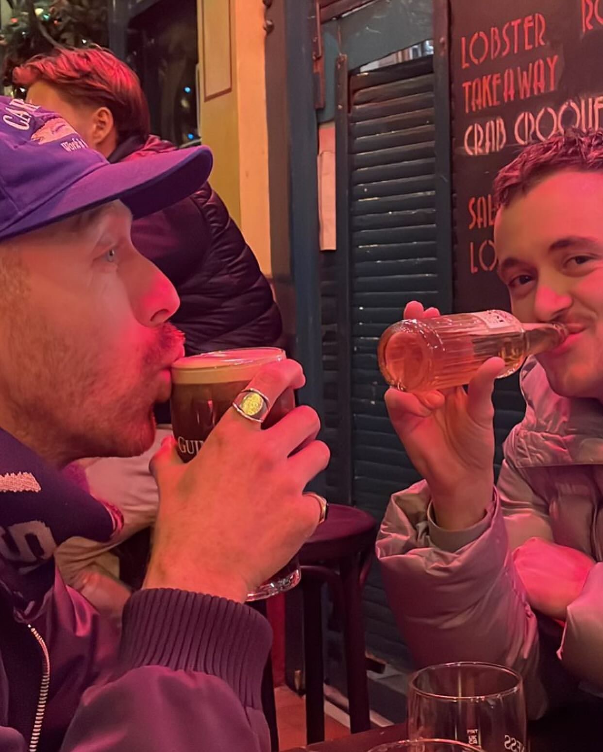 Two men in a bar drinking from a beer glass and a bottle, with visible signage and a green-painted wall in the background.
