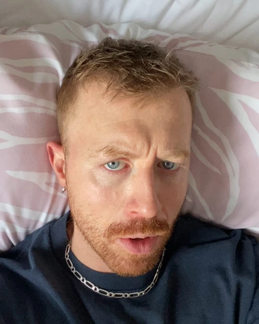 Adult male selfie in bed with a white and pink patterned pillow, wearing a navy shirt and silver necklace with a concerned facial expression.