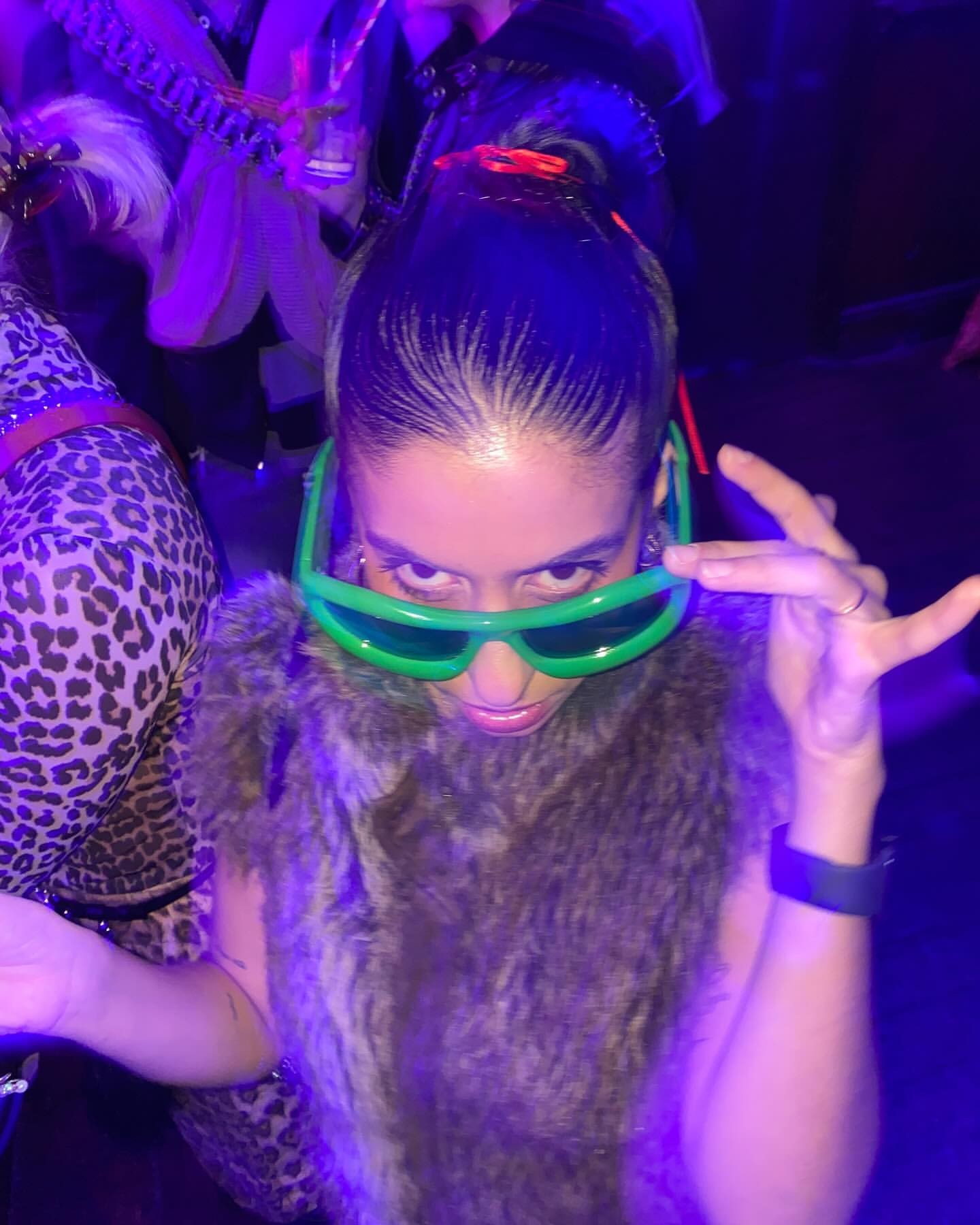 A woman smiles at the camera, wearing a fur vest and oversized green glasses at an event with feathers and leopard print clothing.