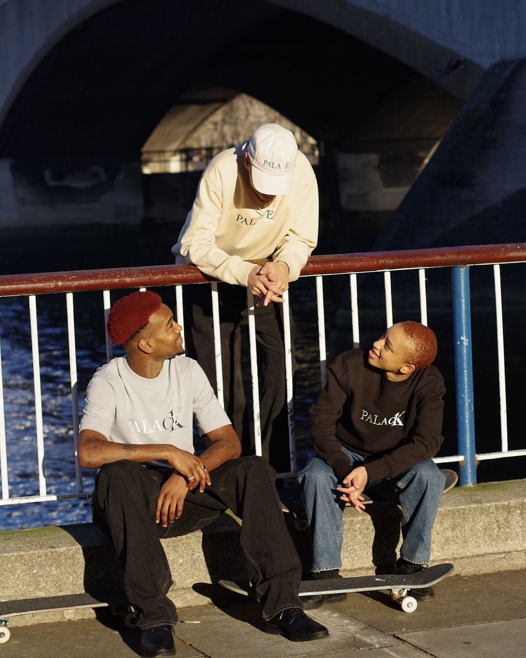 Three young adults in casual attire near a bridge and waterway, two seated on skateboards, all wearing Palace-branded apparel.