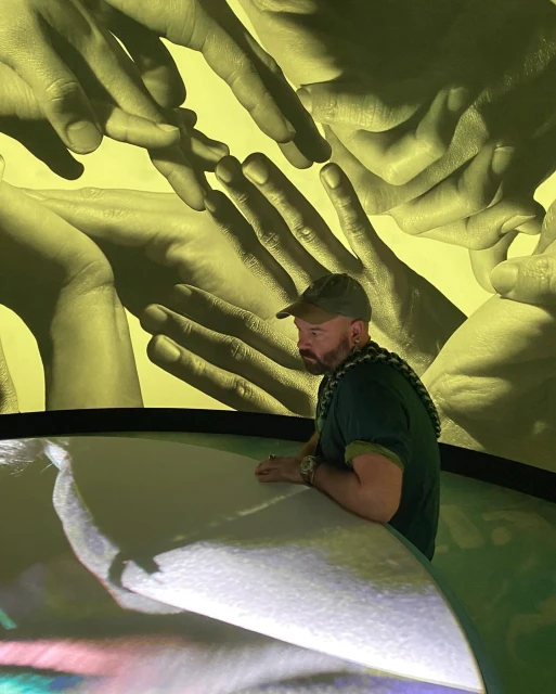 Bearded man stands next to a luminous table, bathed in yellow light from overhead hand projections, creating an immersive art installation.