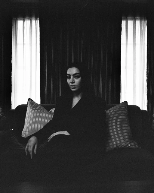 Monochrome portrait of a young woman sitting on a couch with striped pillows and a dark, striped wall with curtained windows in the background.