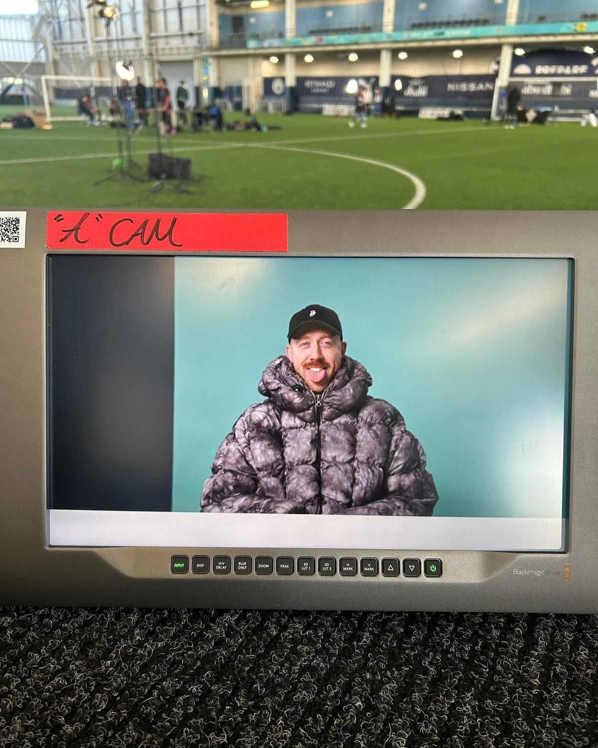 A man is visible on a monitor screen with a green indoor soccer field, lights, and people visible in the distant background.