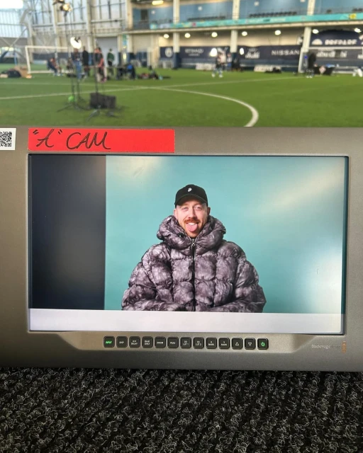 A man is visible on a monitor screen with a green indoor soccer field, lights, and people visible in the distant background.