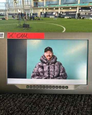A man is visible on a monitor screen with a green indoor soccer field, lights, and people visible in the distant background.