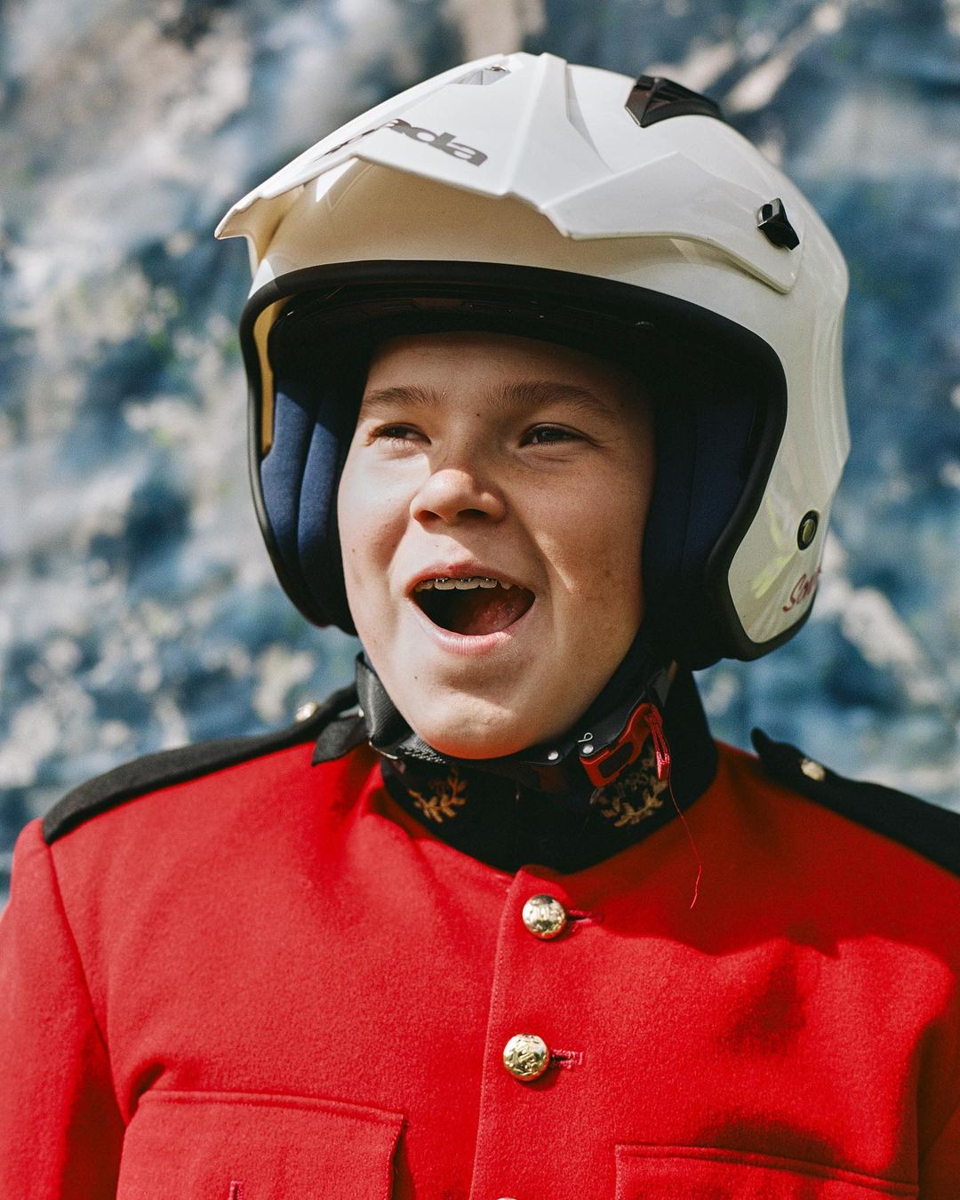 Young boy wearing a red Mountie uniform and white motocross helmet smiles against a blurred background, showcasing an unexpected mix.